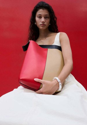 Mujer con vestido blanco sosteniendo un gran bolso rojo y beige sobre un fondo rojo, lleva una pulsera transparente de cuentas.