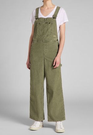 Personne portant une salopette large vert olive avec une poche avant et un t-shirt blanc, debout sur un fond gris avec des baskets blanches.