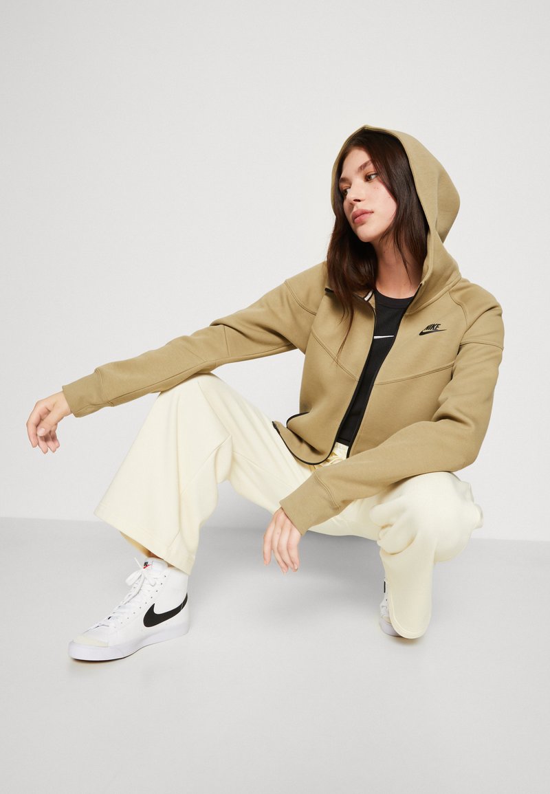 Khakifarbene Zip-Hoodie mit taillierter Passform, langen Ärmeln und einer verstellbaren Kapuze. Kombiniert mit lockeren cremefarbenen Hose und weißen Sneakern.