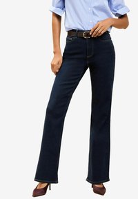 Jean évasé bleu foncé avec une taille haute, des coutures contrastées et une ceinture marron. Associé à une chemise rayée bleu clair et des chaussures à talons.
