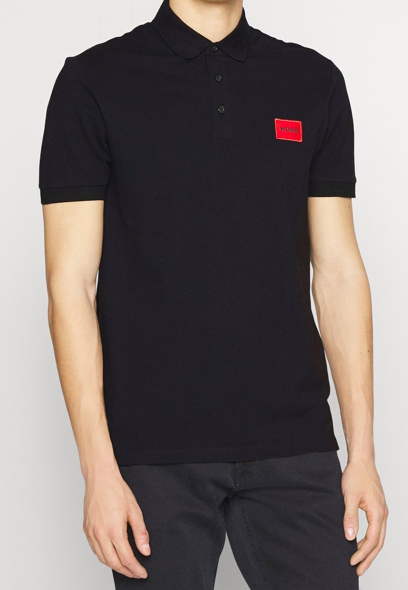 Personne portant un polo noir à manches courtes avec un écusson logo rectangulaire rouge sur la poitrine gauche, associé à un pantalon noir.