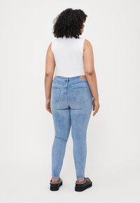 Light blue skinny jeans with back pockets, paired with a white sleeveless top. Back view shows curved seams and a branded waist tag.