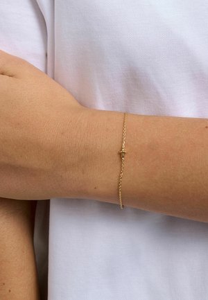 Wrist with light skin wearing a delicate gold chain bracelet featuring a small cross charm against a white fabric background.
