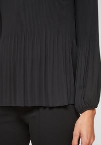 Svart långärmad blus med räfflad textur på den övre delen och samlad detalj vid midjan, med elastiska manschetter och draperad tyg.