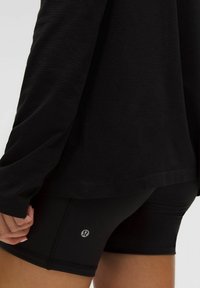Schwarzes Langarmshirt mit lockerer Passform, kombiniert mit schwarzen, taillierten Shorts mit kleinem Logo. Glatte Textur, minimalistisches Design.