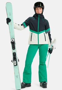 L'équipement de ski comprend une veste noire et crème avec des accents verts, un pantalon vert, un ski et des bâtons. Il présente un matériau imperméable et un design épuré.