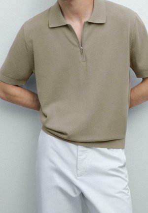 Man draagt een beige kortärmelig gebreide polo met rits aan de voorkant en witte broek, staand tegen een eenvoudige lichte muur.