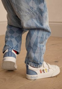 Weiße Sneakers mit blauen Akzenten, ausgestattet mit einem Klettverschluss und Cartoonfiguren an der Seite, kombiniert mit blauen gemusterten Jeanshosen.