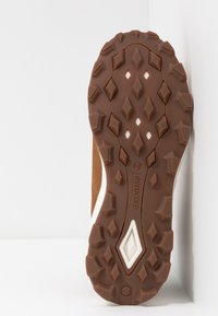 Brun gummisula med ett diamantmönster, prydd med vita accenter och en upphöjd logotyp. Robust konstruktion för bra grepp på olika ytor.