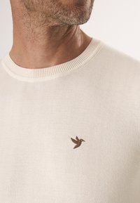 Jasno beżowy sweter z dzianiny, z okrągłym dekoltem i małym haftem brązowego ptaka na piersi. Miękka faktura i swobodny styl.