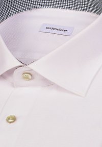 Gros plan sur le col d'une chemise blanche et les boutons supérieurs montrant l’étiquette de la marque Seidensticker à l’intérieur.