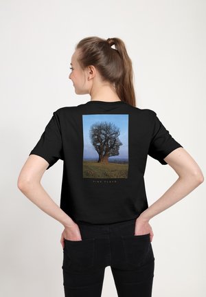 Ung kvinde med hænder i lommerne bærer en sort T-shirt med et træ formet som en profil af et ansigt med teksten Pink Floyd.