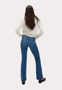Mujer con cabello largo castaño que lleva un suéter crema, jeans acampanados azules y zapatos negros, de pie con las manos en las caderas, mirando hacia atrás.