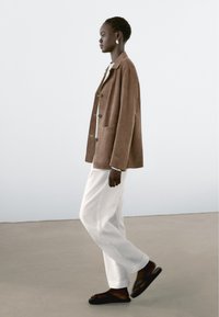 Braune Wildleder-Übergroßjacke mit silbernen Knöpfen, getragen über einem weißen Oberteil und weißen Hosen. Schwarze Flip-Flops vervollständigen das Outfit.