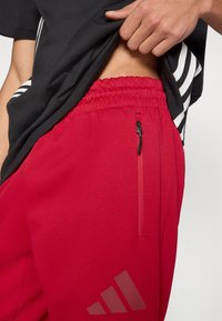 Κόκκινο αθλητικό παντελόνι με ελαστική μέση, μία τσέπη με φερμουάρ και διακριτικό λογότυπο. Το υλικό φαίνεται λείο και ελαφρύ.