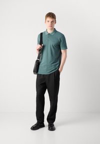 Grønn polo med krage og tre knapper, kombinert med svarte bukser. En svart budveske og svarte sko fullfører antrekket.