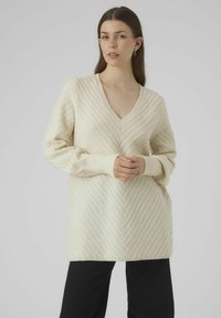 V-neck knit sweater in light cream with textured chevron pattern; relaxed fit with ribbed cuffs and hem. Paired with black pants.