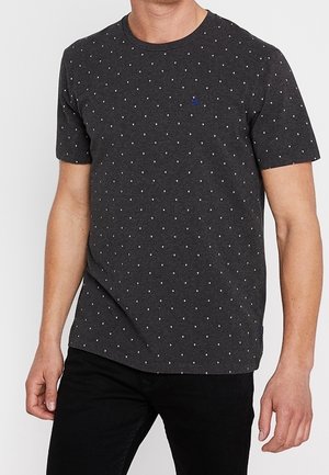 Mężczyzna ubrany w ciemnoszary t-shirt z krótkim rękawem w małe białe kropki i czarne spodnie, stoi z rozluźnionymi ramionami.
