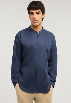 Junger Mann mit dunklen lockigen Haaren, der ein marineblaues Hemd mit Stehkragen und helle beige Hose vor einem schlichten Hintergrund trägt.