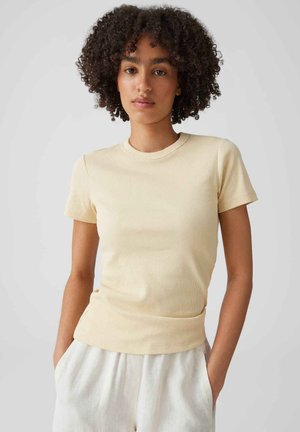 Femme aux cheveux bouclés portant un haut côtelé beige à manches courtes et un pantalon clair, les mains dans les poches, sur un fond uni.