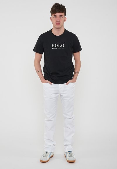 Maglietta nera in cotone con logo "POLO RALPH LAUREN", abbinata a pantaloni in denim bianchi e sneakers beige/blu, in piedi su uno sfondo bianco.