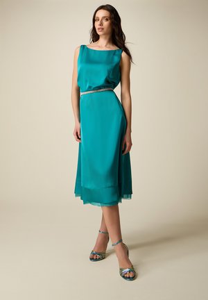 Mujer de pie con vestido midi de satén color verde azulado sin mangas, cinturón plateado y sandalias de tacón alto a juego con tiras, sobre fondo beige.