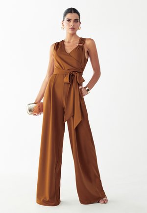 Kobieta ubrana w bezrękawnikowy kombinezon w kolorze rdzawego z szerokimi nogawkami i paskiem w talii, trzymająca złotą kopertówkę oraz nosząca złote kolczyki i bransoletkę.