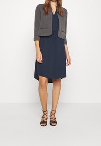 Laivastonsininen mekko, jossa on nyörivyötärö ja A-linjainen leikkaus, yhdistettynä lyhyeen harmaaseen bleiseriin ja mustiin narusaappaisiin. Sujuva kangas, istuva malli.