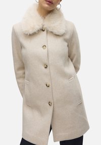 Manteau en laine beige avec un col en fausse fourrure, six boutons sur le devant et deux poches latérales. Le tissu présente une apparence douce et texturée.
