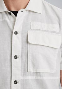 Camisa blanca de manga corta hecha de tejido texturizado, con botones negros, un bolsillo en el pecho y una etiqueta en relieve con "PME LEGEND."