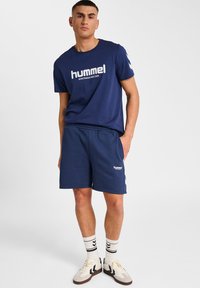 Ung man som står i navyblå hummel sportkläder med t-shirt och shorts, vita strumpor och vita sneakers mot en ren vit bakgrund.