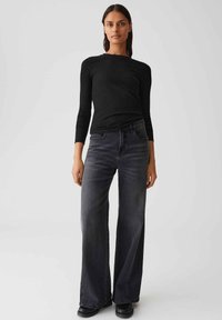 Femme portant un haut noir ajusté à manches longues en côtes et un jean noir délavé taille haute à jambes larges, se tenant devant un fond uni.