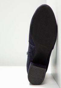 Semelle noire d'une botte en daim bleu marine foncé avec talon et semelle extérieure en caoutchouc texturé, présentée verticalement contre une surface et un mur blancs.