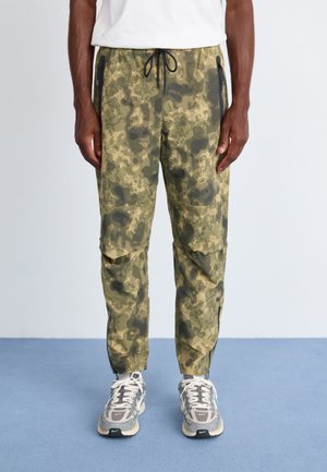 Camo-patterned cargo pants in olive green with zippered pockets, elastic waistband, and tapered legs. Durable fabric with a textured finish.