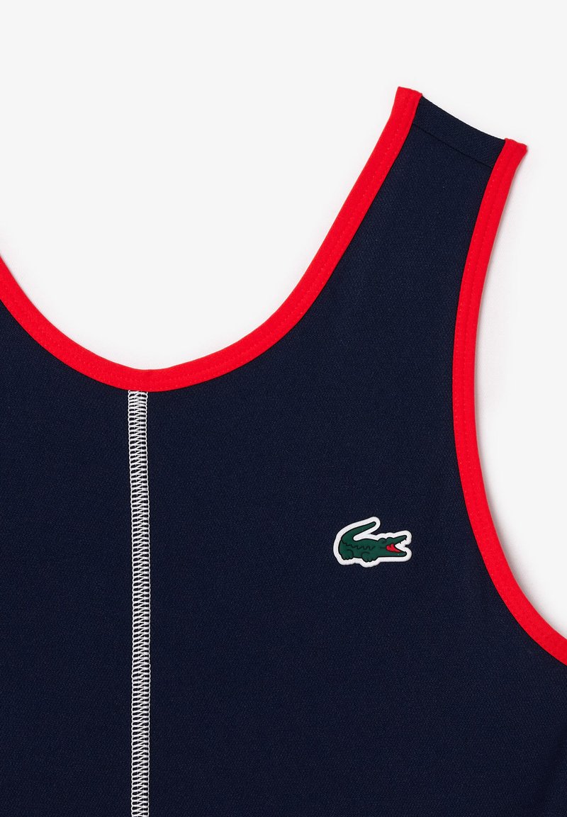 Marineblauw tanktop met rode details, inclusief een ronde halslijn. Voorzien van een witte stikselslijn en een merklogo van een groene krokodil aan de onderzijde links.