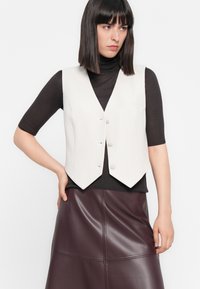 Chaleco blanco con cuello en V, cuatro botones y un único bolsillo en el pecho, superpuesto a una blusa negra de canalé, combinado con una falda de cuero burdeos.