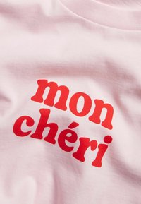 Rosa bomulls-t-shirt med djärv röd text som säger "mon chéri." Texten har ett runt, lekfullt typsnitt och en mjuk textur.