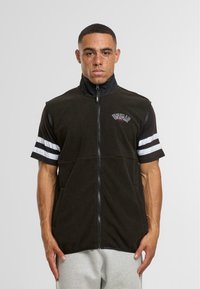 Gilet en polaire noir avec col haut, zip complet et logo. Présente des manches courtes avec des rayures blanches. Texture douce et deux poches latérales.