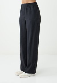 Weite Hosen aus glattem, dunklem Stoff mit elastischem Bund, die einen lockeren Sitz haben und mit weißen Sneakers kombiniert sind.