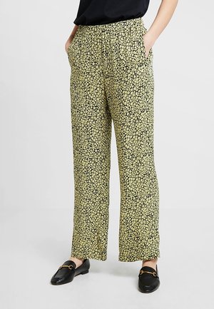 Frau trägt lockere gelbe Hose mit kleinem schwarzem Blumenmuster, schwarzen Oberteil und schwarzen Slippern mit goldenen Details, steht mit Händen in den Taschen.