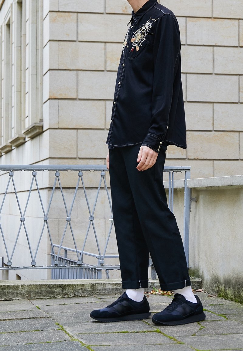 Schwarzes Hemd mit Knöpfen und bestickten Details, kombiniert mit schwarzen, kappe Hosen und schwarzen Sneakers mit weißen Socken, vor einer Steinmauer.