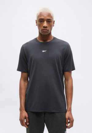 Νεαρός με κοντό ξανθό μαλλί, φορώντας ένα μονόχρωμο σκουρόγυαλό γκρι T-shirt της Reebok και αντίστοιχο σκούρο παντελόνι, στέκεται μπροστά σε ένα απλό φωτεινό φόντο.