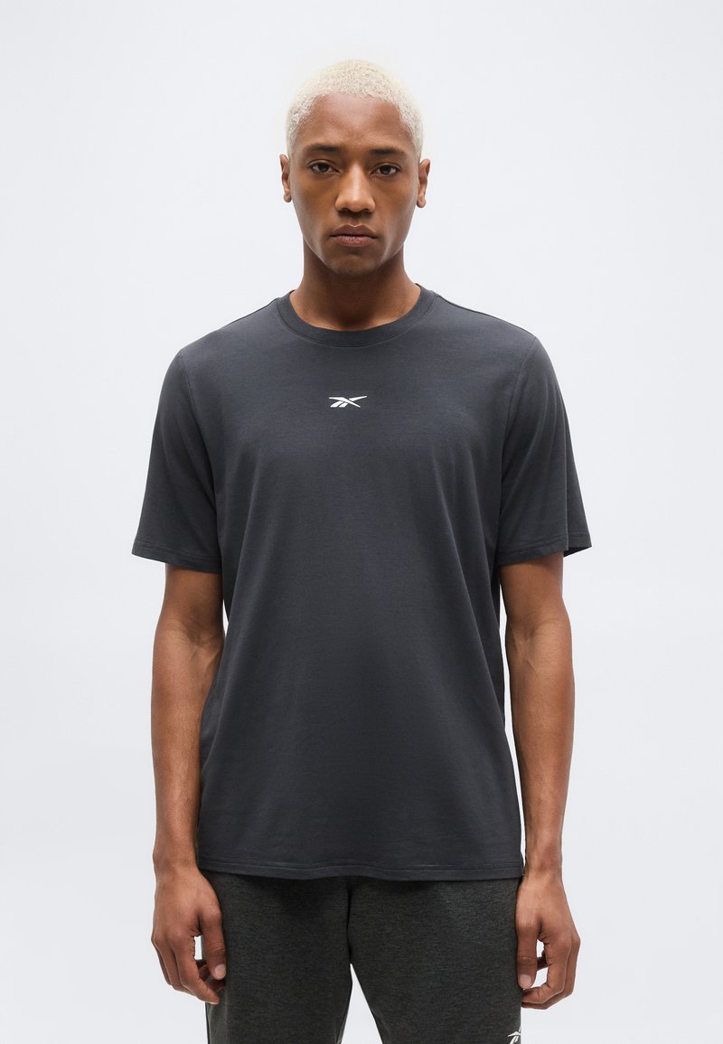 Νεαρός με κοντό ξανθό μαλλί, φορώντας ένα μονόχρωμο σκουρόγυαλό γκρι T-shirt της Reebok και αντίστοιχο σκούρο παντελόνι, στέκεται μπροστά σε ένα απλό φωτεινό φόντο.