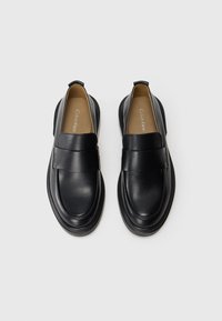 Pantofi negri din piele, tip loafers, cu talpă groasă și curele late, branduți Calvin Klein în interior, văzuți dintr-o perspectivă de sus pe un fundal alb.