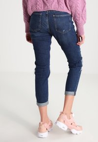 Tmavě modré džíny s tapered střihem, ohrnutými rukávy a oprýskanými detaily, doplněné světle růžovými teniskami a texturovaným růžovým svetr.