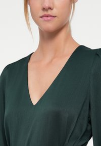 Dark green dress featuring a deep V-neck, textured fabric with subtle vertical stripes, and softly puffed sleeves.