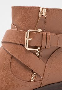 Bottine en cuir synthétique marron avec une sangle croisée ornée d'une boucle dorée et une fermeture éclair sur le côté. Surface texturée, bout arrondi.