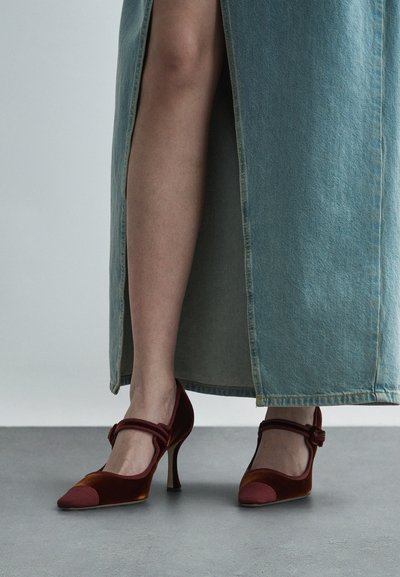 Röda och vinröda sammetshöga klackskor med spetsig tå och ankelrem, i kombination med en blå denimkjol med slits.