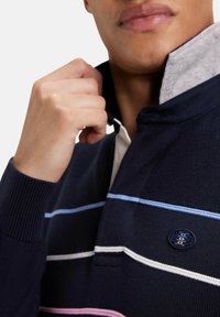Polo marine avec des rayures horizontales bleu clair et blanches. Il présente un col intérieur gris contrastant et une patch logo sur la zone de la poitrine.