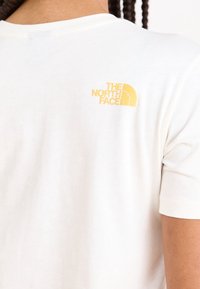 Camiseta blanca de algodón con un pequeño logo amarillo bordado en la parte superior de la espalda, cuello redondo y mangas cortas.